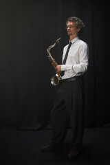 Naklejka premium Vertical studio shot of mature male musician dressed in formal concert attire posing with saxophone in hands standing against black curtain, copy space