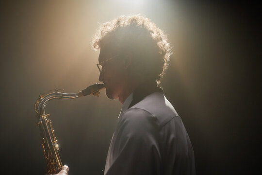 Side view shot with backlight of adult curly man improvising on saxophone playing enchanting jazz music under spotlight on stage, glare effect, copy space