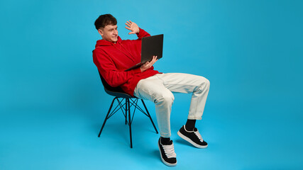 Relaxed young man on chair waving to laptop screen during online conversation, against blue background. Concept of distance work, remote calls, digital talk, emotions, casual communication, style.