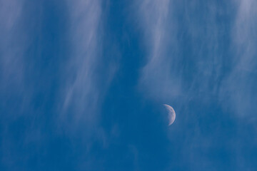 A faint crescent moon appears through delicate, wispy clouds in a tranquil blue sky, creating a...