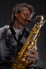 Fototapeta premium Vertical dynamic shot of professional male saxophonist in glasses playing jazz music with devotion against grey background in studio with backlight, slow shutter effect