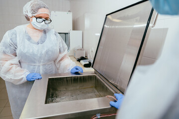 Lab technician in protective suit checking water level and temperature in open breast milk pasteurizer. Sterilization and hygiene protocols followed.
