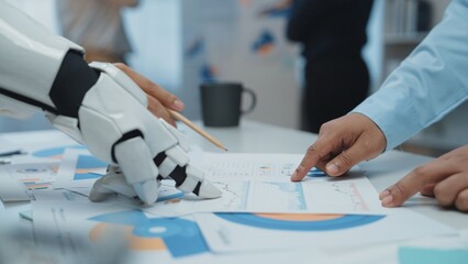 A robot hand collaborates with a human hand over charts and documents in a modern office, showcasing the integration of technology in business strategies and analysis.Noogenesis