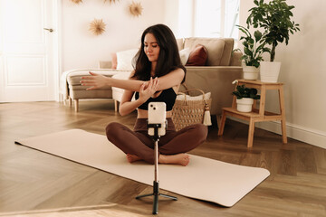 A fitness woman does sports at home using a mobile phone and a tripod while sitting on a exercise mat in a cozy interior. The influencer records an online workout. Healthy lifestyle. Selective focus