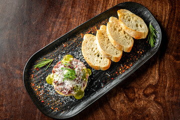 A gourmet dish featuring finely chopped meat topped with grated cheese and herbs, served alongside slices of crusty bread on a dark plate, with a sprinkle of spices.