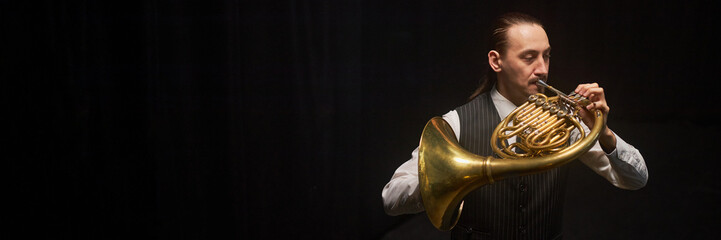 Website header shot of male French horn player concentrated on performing jazz music on while standing in bright light on stage against black backdrop, copy space