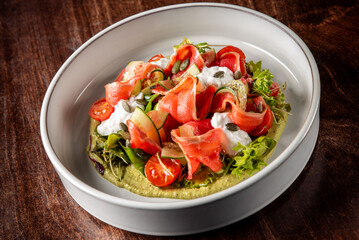 A vibrant salad featuring thinly sliced vegetables, including tomatoes and greens, drizzled with creamy dressing and garnished with seeds, presented in a white bowl on a wooden surface.
