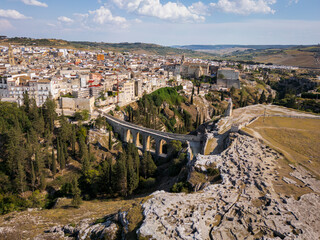 Scenic view of Gravina in Puglia, Italy, showcasing historic stone architecture, the iconic Roman bridge over the ravine, and the picturesque old town under a clear blue sky.