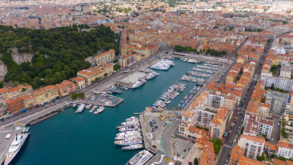 Drone view of Nice, France, showcasing the vibrant harbor filled with yachts and boats, surrounded by colorful historic buildings and the stunning coastline of the French Riviera under a blue sky. 