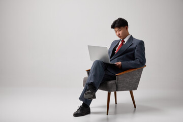 Confident Asian man sitting in chair and working or browsing in laptop against grey studio...