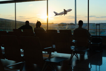 backlit image of people waiting in an airport lounge while watching a plane take off through a...