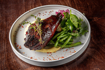 A succulent grilled meat portion served on a bed of creamy mashed potatoes, accompanied by green beans and a fresh herb salad, all elegantly plated on a white dish with a rustic wooden background.