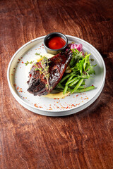 A succulent grilled meat portion served on a bed of creamy mashed potatoes, accompanied by green beans and a fresh herb salad, all elegantly plated on a white dish with a rustic wooden background.