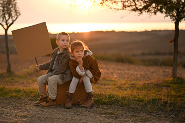 Fototapeta premium Boy and girl on a rural road at sunset. sits on a suitcase, waiting for a ride or bus. A nostalgic scene of childhood, wanderlust, and adventure in the warm evening light.