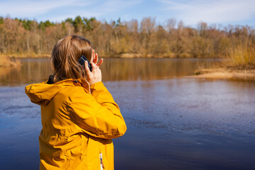 A young woman in a yellow jacket makes an emotional phone call by the river, appearing distressed....
