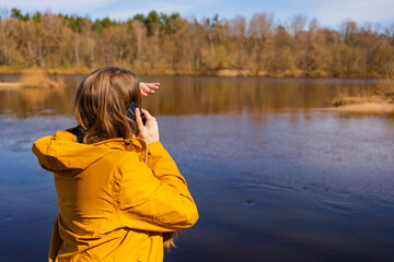 A young woman in a yellow jacket makes an emotional phone call by the river, appearing distressed. The situation seems urgent or personal as she seeks help or shares critical news.
