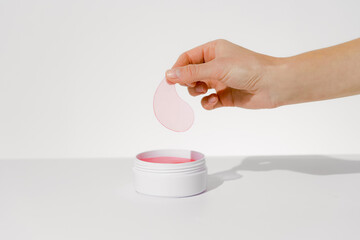 Female hand holding pink hydrogel patch over jar of patches on white isolated background. Concept of beauty products, skin care under eyes, reduction of dark circles and wrinkles.