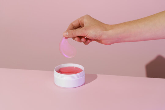 Female hand holding pink hydrogel eye patch over white jar with patches on pink isolated background. Concept of beauty products for skin care under eyes, reducing dark circles and wrinkles.