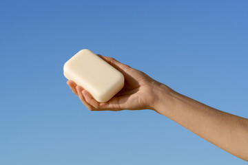 Female hand holding a creamy bar of soap against a blue sky background. Concept of cleanliness,...