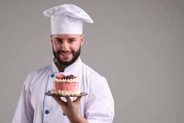 Happy confectioner in uniform holding delicious cake with berries on light grey background