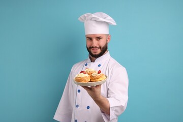 Happy confectioner in uniform holding delicious profiteroles with strawberries on light blue background