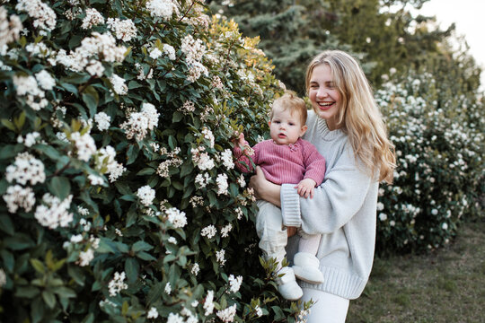 Happy stylish mother holding baby girl 1 year old wearing knitted sweaters posing with flowers over nature background close up. Motherhood. Springtime.