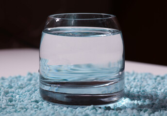 A transparent glass filled with clean water standing on a light surface covered with small decorative blue pebbles. 