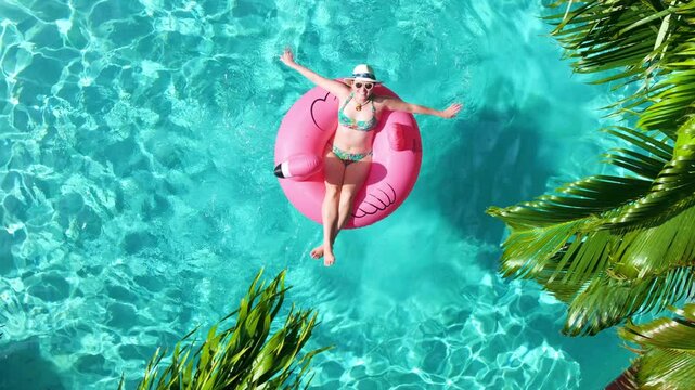 Aerial shot of a tropical backyard pool with a woman on a flamingo float, palm trees casting shadows over clear turquoise water, colorful loungers and a striped parasol creating perfect vacation vibe.