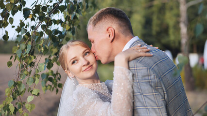 Groom gently kissing bride's cheek beneath birch branches, wedding day radiating intimate love and...