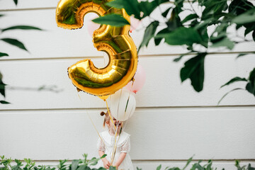 Little girl celebrates her third birthday with colorful balloons near a white wall