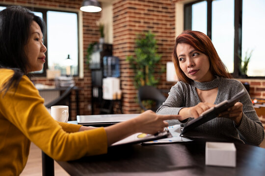 Businesswomen collaborate, exchanging ideas as one listens to the strategy presentation of her colleague during meeting. Asian entrepreneurs discussing startup plans, holding clipboards in office.