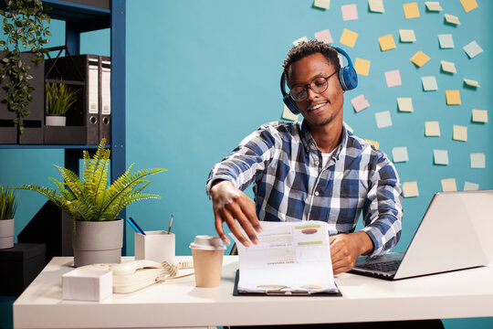 Calm male accountant listening to music and going over company documentation. Business advisor wearing headphones and flipping through papers on clipboard, reviewing financial data in marketing firm.