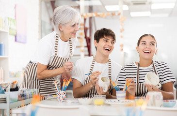 Friendly older mother and young adult daughter and son, wearing aprons, talking cheerfully while working on painting ceramic dishware in family pottery workshop. Generational collaboration concept..