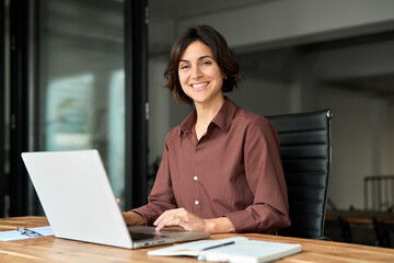 Portrait of confident businesswoman leader in her 30s at work desk. Smiling Hispanic young woman entrepreneur, happy female executive manager looking at camera sitting at work with laptop.
