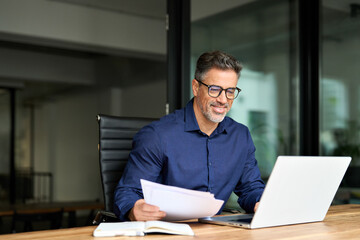 Smiling man executive manager or business owner of middle age reading paper working on computer....