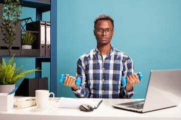 African american businessman seated at office desk, doing arms workout with dumbbells. Active young...
