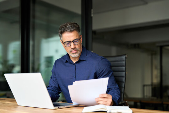 Middle aged professional business man account bank manager using laptop computer reading document at work. Busy mature male executive working on laptop checking paper financial report in office.