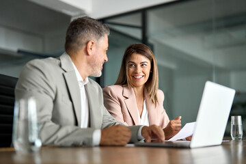 Two mid aged colleagues company board discussing digital strategy at corporate meeting. Professional business executive managers office team working using laptop computer sitting at table. Candid shot