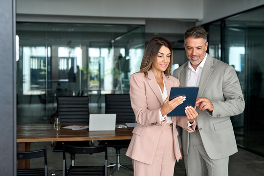 Two busy older professional corporate business executives man and woman wearing suits working on digital project plan holding tablet technology device having discussion standing in office at meeting.