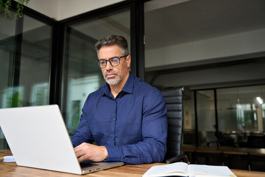 Busy serious middle aged man executive business owner or investor using laptop. Middle aged professional businessman ceo manager wearing glasses sitting at desk working looking at computer in office.