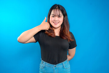 Fototapeta premium Portrait of a cheerful young woman smiling and giving a thumbs-up gesture. Confident and positive expression against a vibrant blue background, ideal for advertising and marketing.