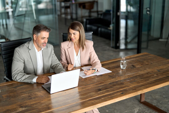 Two professional business executive managers office team working using laptop computer sitting at table. Happy older colleagues discussing company digital strategy at corporate meeting. Authentic shot