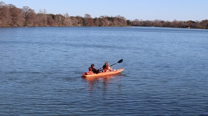 Two girls kayak on a calm lake in an orange vessel, surrounded by bare trees under a clear blue...