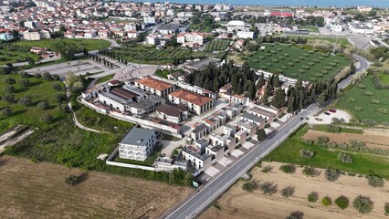 This aerial view captures a modern Italian cemetery, featuring an array of distinct mausoleums and tombs nestled among cypress trees, revealing organized pathways and a solemn, architectural landscape