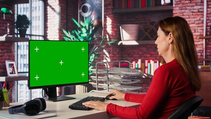 Adult woman typing on pc with chroma key screen at her loft desk, engaging in online work,...