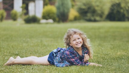 Fototapeta premium Happy young girl with curly blonde hair relaxing on a green lawn, enjoying a sunny family day in the backyard