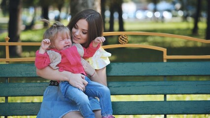 Caring mother soothing distressed child, seated on park bench under bright sunlight