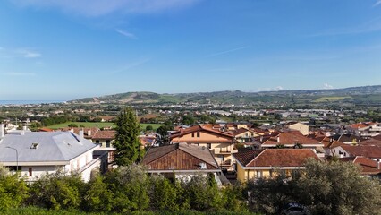 Fototapeta premium A sunlit panoramic view of the Italian town of Roseto Degli Abruzzi, showing red-tiled roofs, a green valley, and a distant coastline under a blue sky.