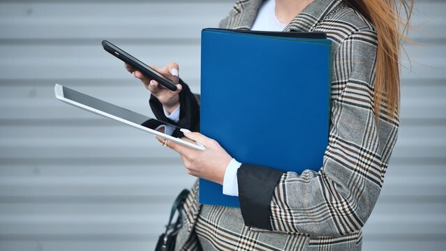 Businesswoman walking and using smartphone and digital tablet while holding a blue folder