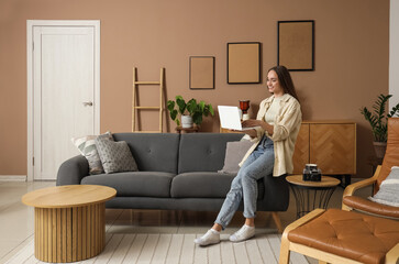 Fototapeta premium Young pretty woman sitting on sofa with laptop in beige living room